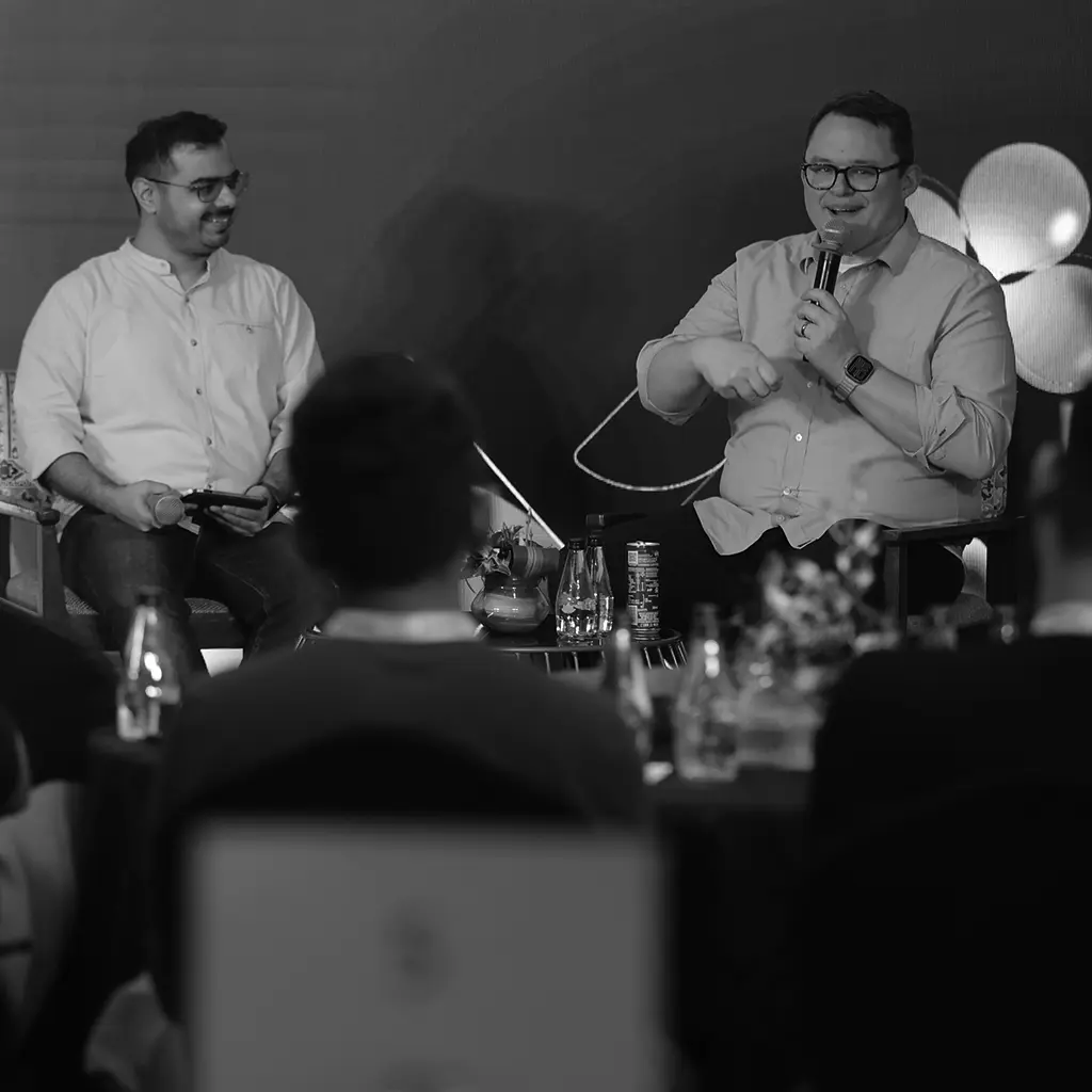 A black-and-white photograph of an indoor event space featuring a staircase with attendees walking and sitting. At the top of the stairs is a large GitHub Octocat logo illuminated in the background. To the right, there is a sign labeled "GitHub Expert Center," inviting attendees to seek assistance or information. The space has a modern industrial design with visible beams and open seating areas.